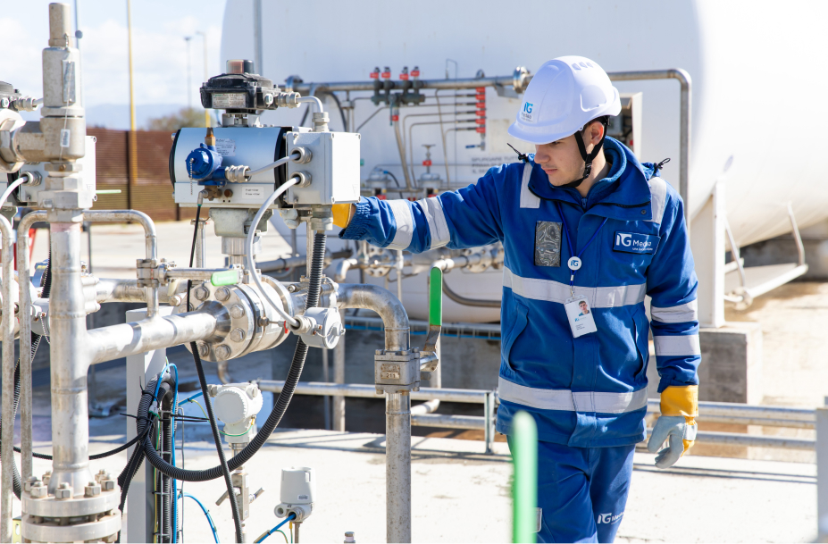 Un tecnico Italgas, in tuta e casco di sicurezza, lavora su una valvola di un impianto industriale, circondato da tubature e strumentazione tecnica.