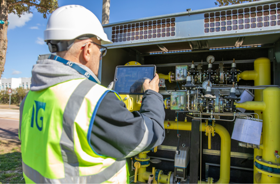 Un tecnico Italgas, con casco e gilet riflettente, utilizza un tablet per monitorare una cabina di regolazione gas, circondato da tubature e strumenti di controllo.