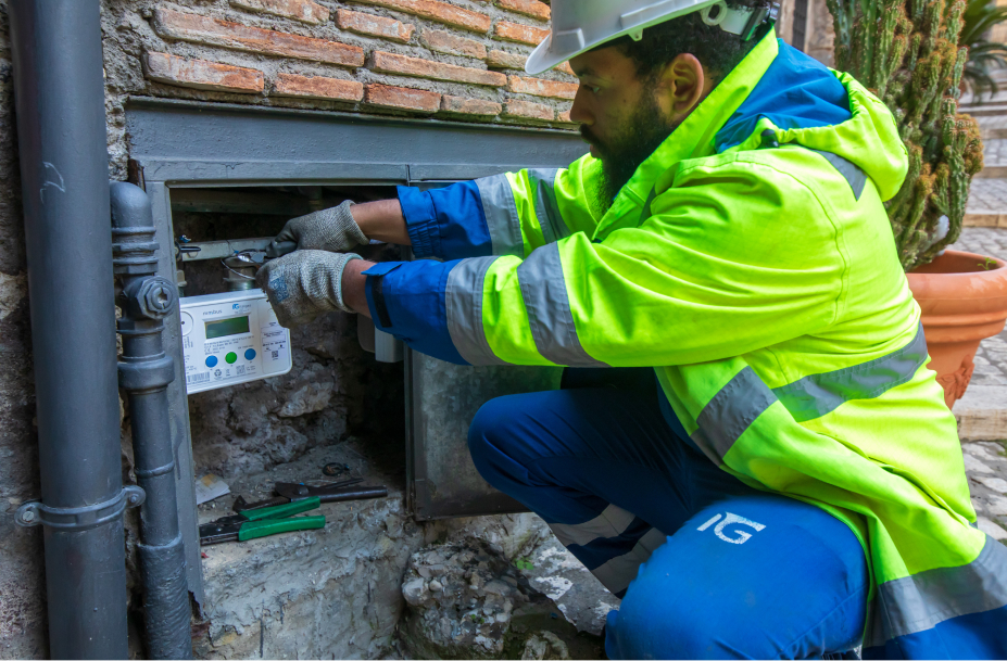 Un operaio Italgas, inginocchiato, installa un contatore Nimbus in una nicchia murata in una strada di ciottoli, lavorando con attrezzi.