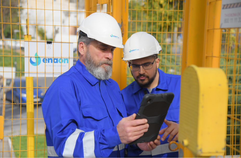 Due tecnici Italgas in tuta e casco osservano un tablet, logo Enaon sullo sfondo.