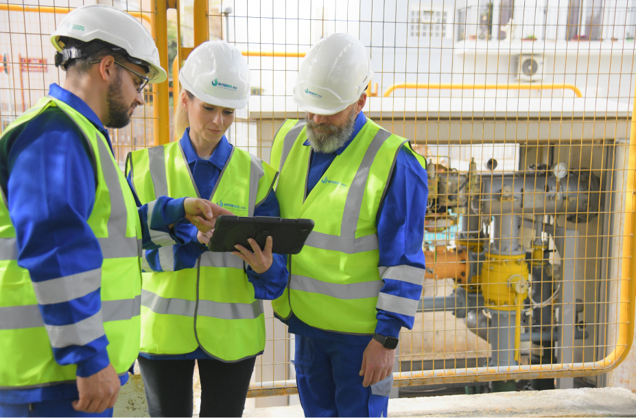 Tre tecnici Italgas in alta visibilità controllano dati su tablet davanti a impianti di rete gas.