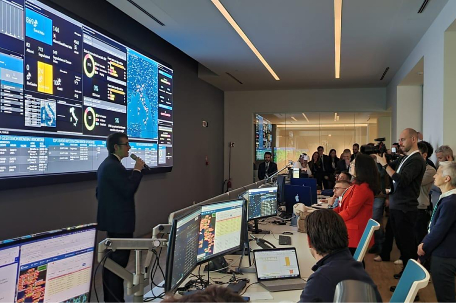 Sala di controllo Italgas con persone che osservano schermi con dati e un uomo che spiega.