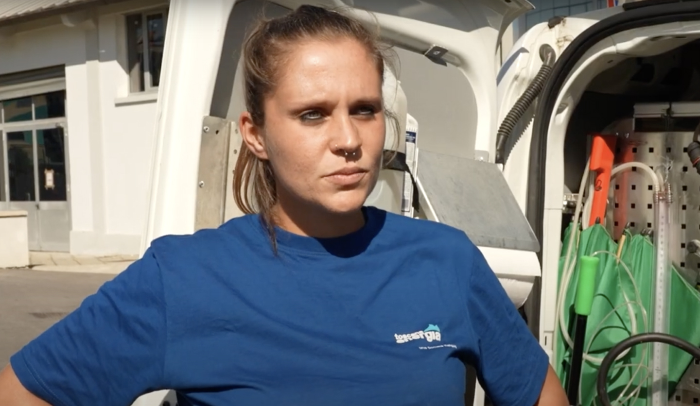 Donna in maglietta blu con logo Italgas, di fronte a veicolo attrezzato, in giornata soleggiata.