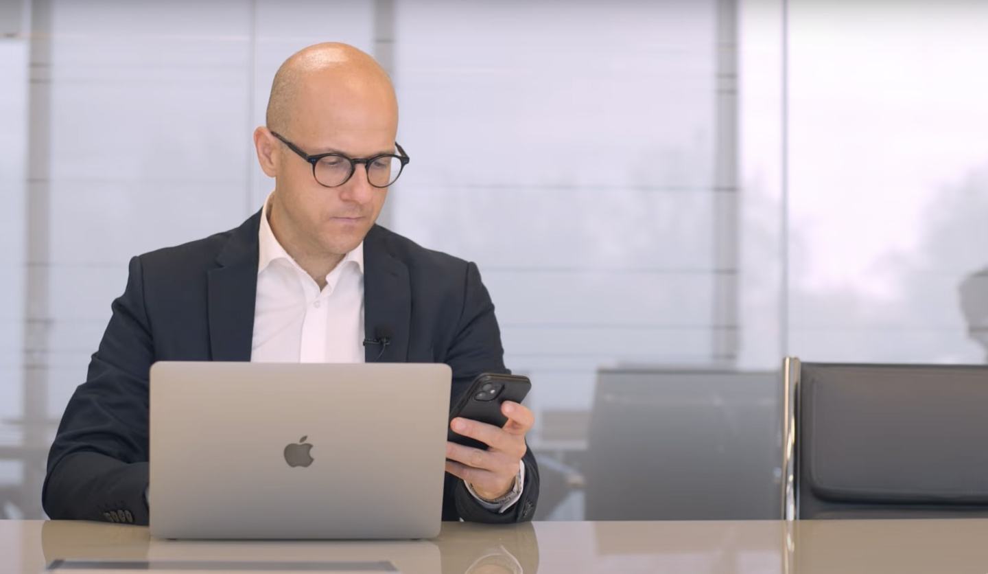 Uomo in abito con occhiali controlla smartphone e laptop in ufficio, simboleggia il controllo dei costi di Italgas.