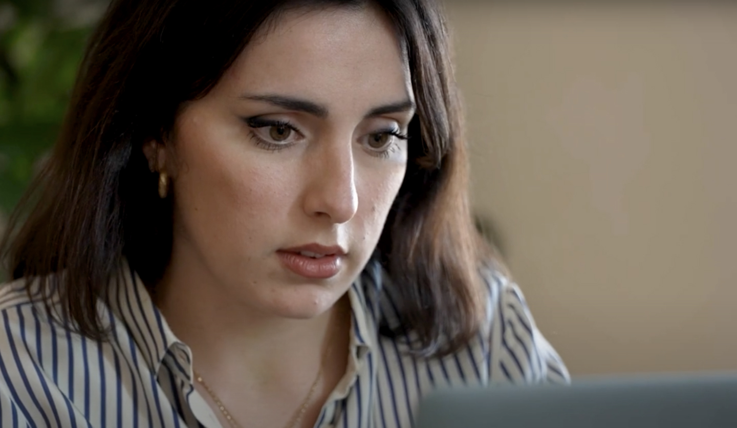 Donna concentrata al computer in ufficio, indossa camicia a righe. Non c'è branding visibile.