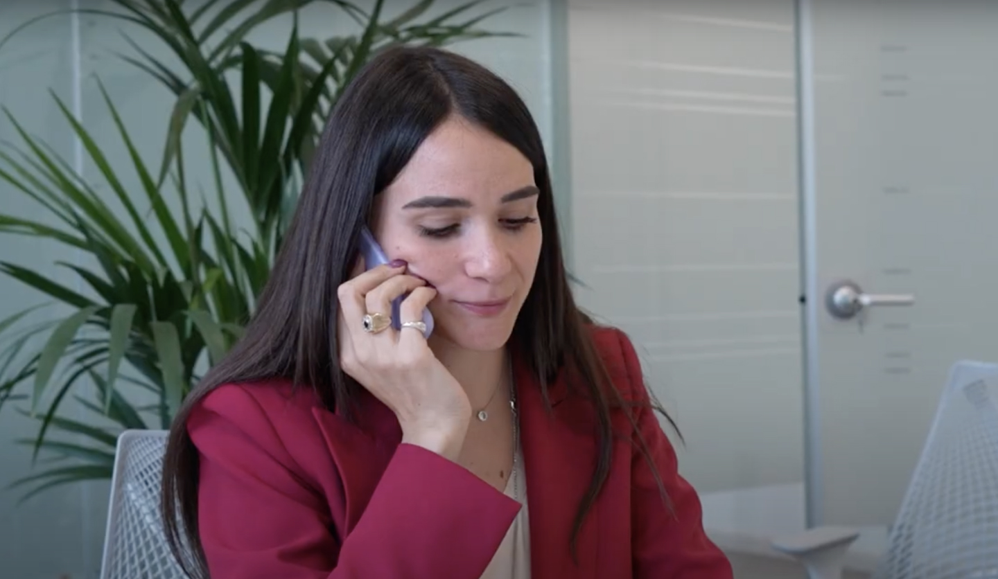 Donna in giacca rossa parla al telefono in un ufficio luminoso con piante verdi sullo sfondo.