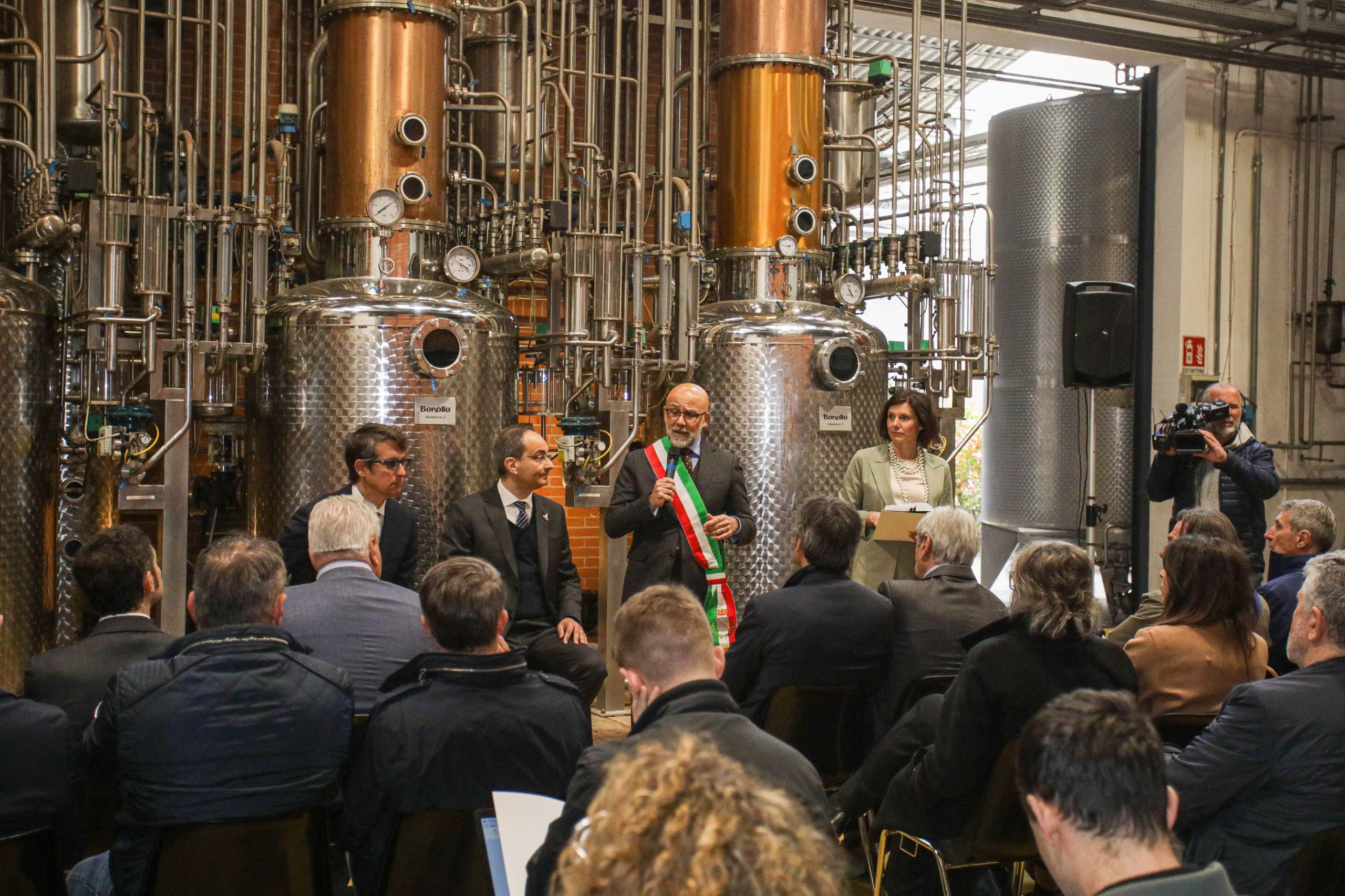 A man wearing a tricolor sash speaks in front of an audience in a grappa industrial facility, surrounded by metal tanks and equipment. Next to him are other speakers and a presenter.