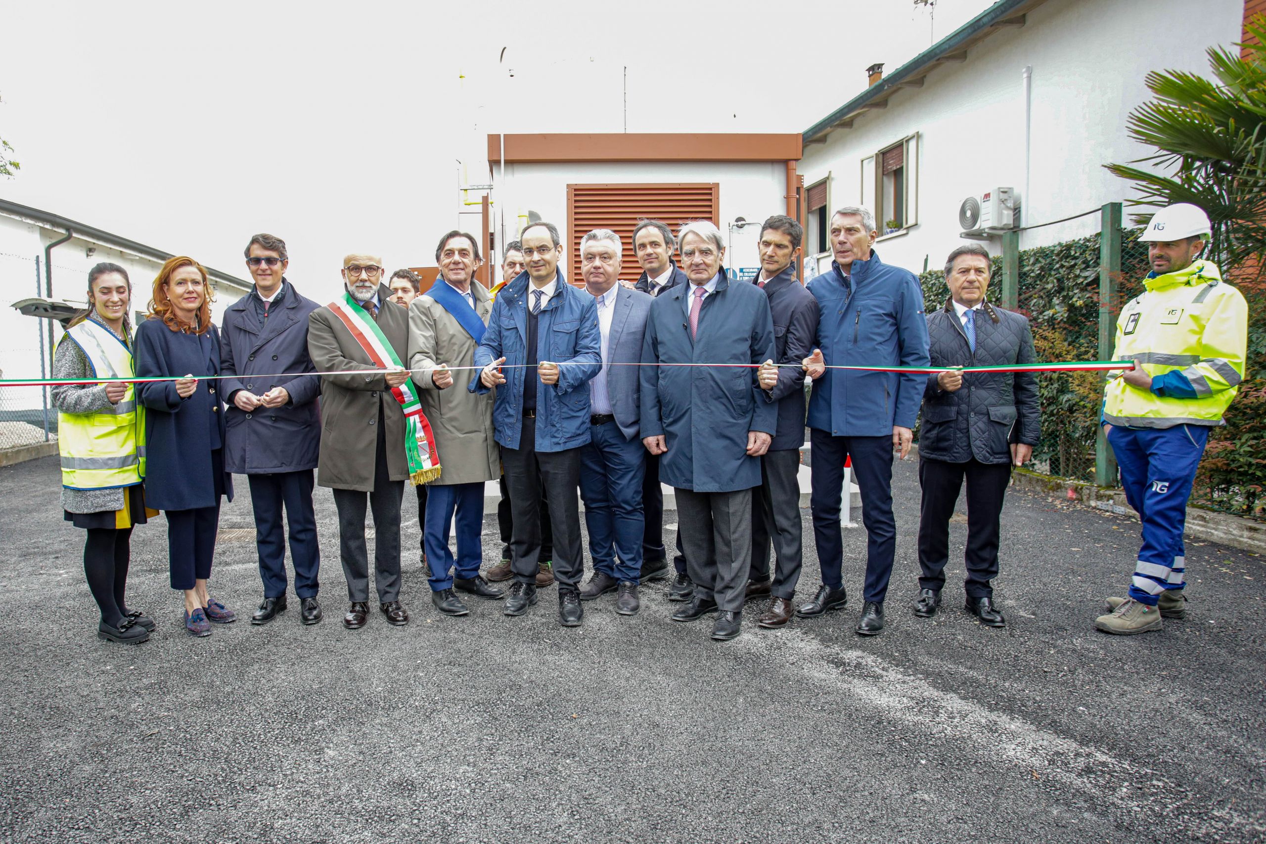 Un gruppo di persone in abiti formali, inclusi ufficiali con fasce tricolori, partecipa a una cerimonia di inaugurazione, tenendo un nastro tricolore per un taglio ufficiale all'aperto.