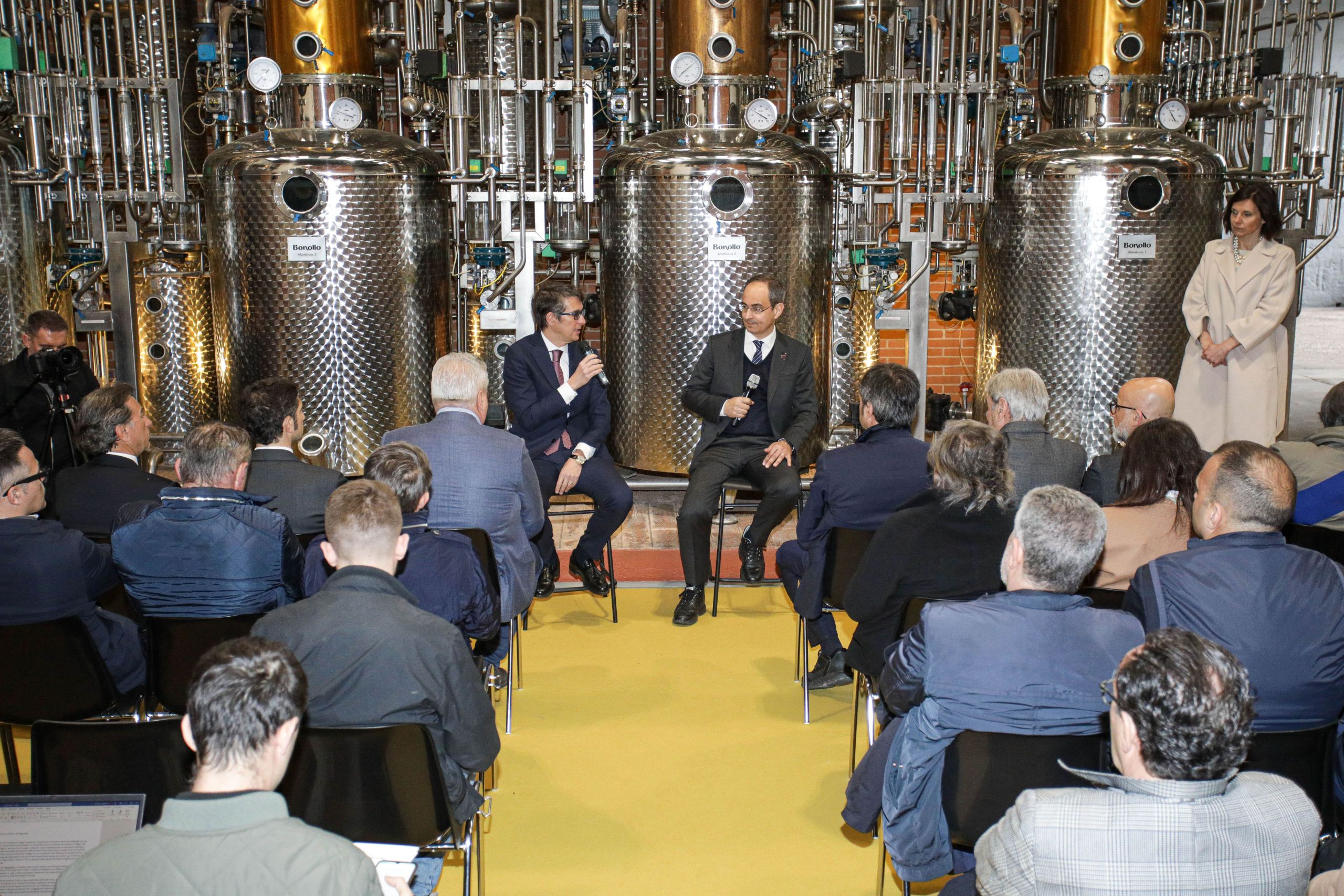 Due uomini in abiti formali parlano con microfoni davanti a un pubblico in un impianto industriale di produzione di grappa, circondati da serbatoi metallici. Un pubblico attento li ascolta seduto.