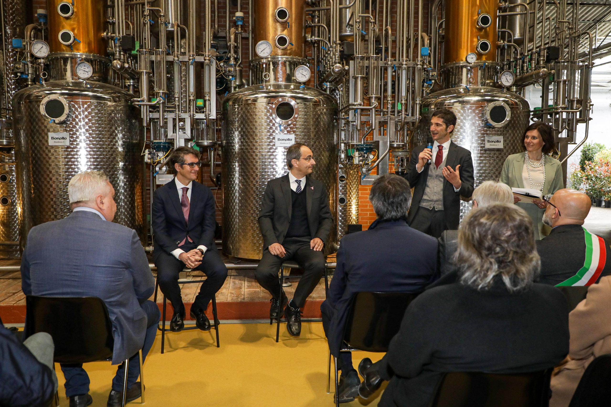 A man in formal attire speaks into a microphone in front of an audience in a grappa industrial facility, while other speakers listen, surrounded by metal equipment.
