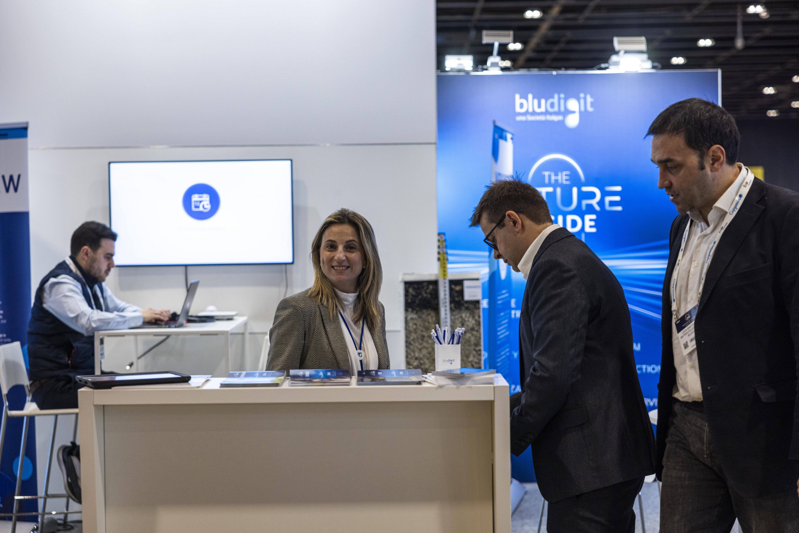 Un evento aziendale con persone a un banco d'accoglienza. Una donna sorride, mentre due uomini leggono materiale promozionale. Sullo sfondo, un uomo lavora al computer.