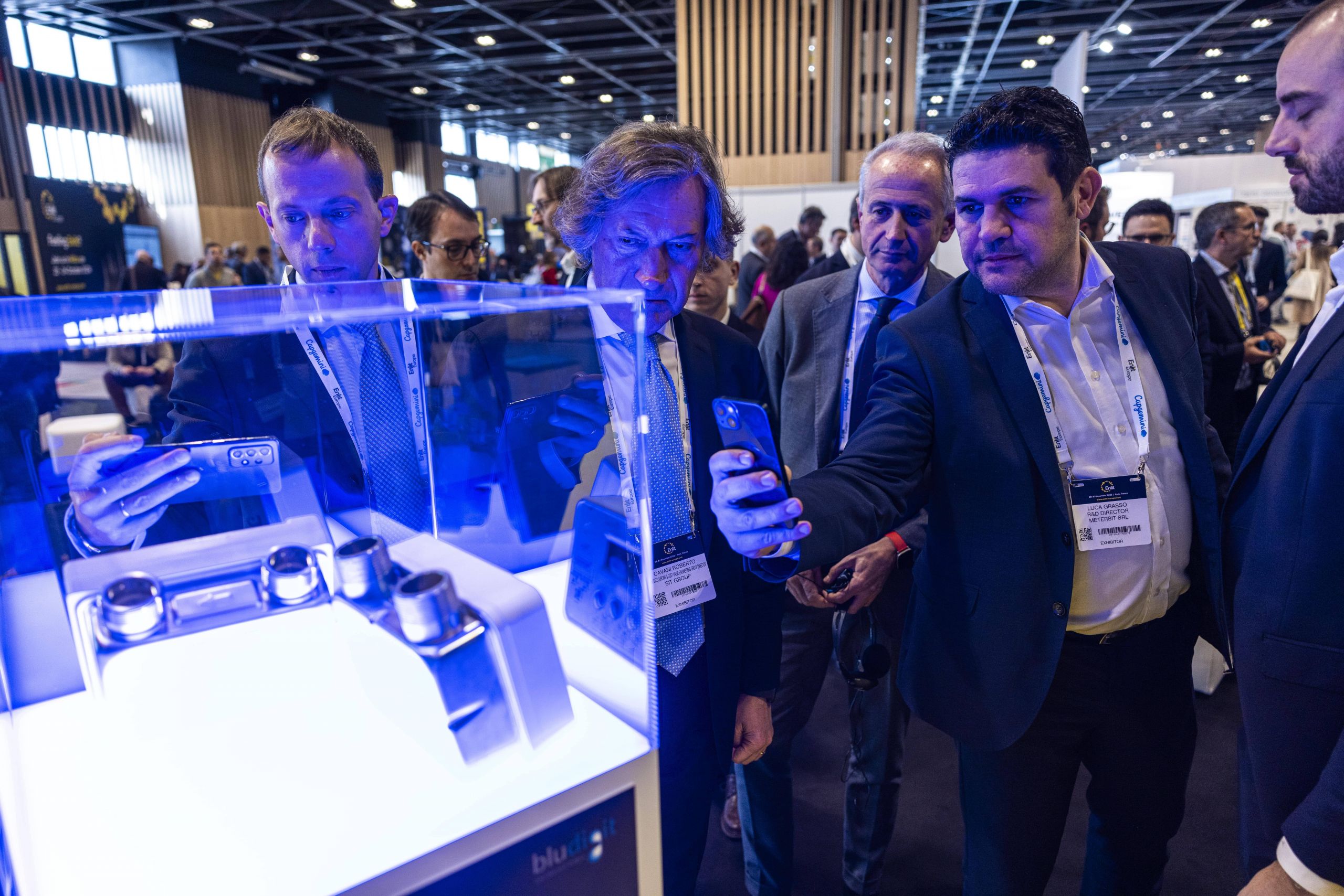 Alcuni uomini in abiti da lavoro osservano e fotografano con i loro telefoni il contatore Nimbus esposto in una teca illuminata di blu, durante un evento professionale in una fiera.