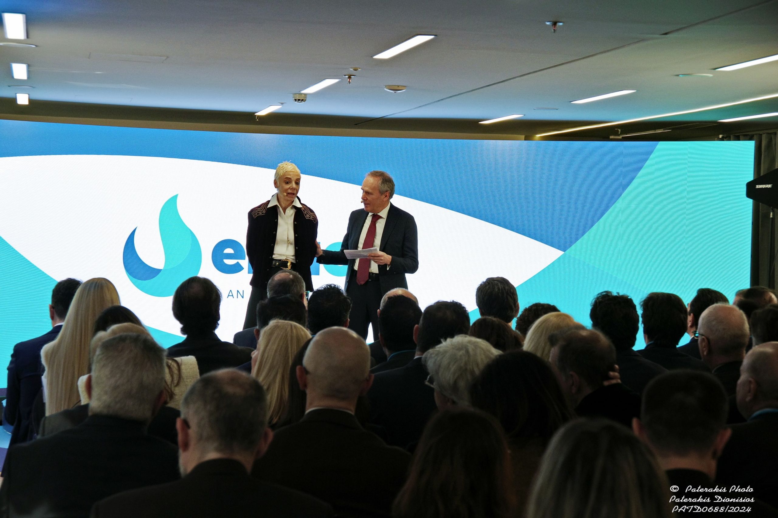 Due persone sul palco durante un evento aziendale con ascoltatori in primo piano e la proiezione del grande logo di Enaon sullo sfondo.