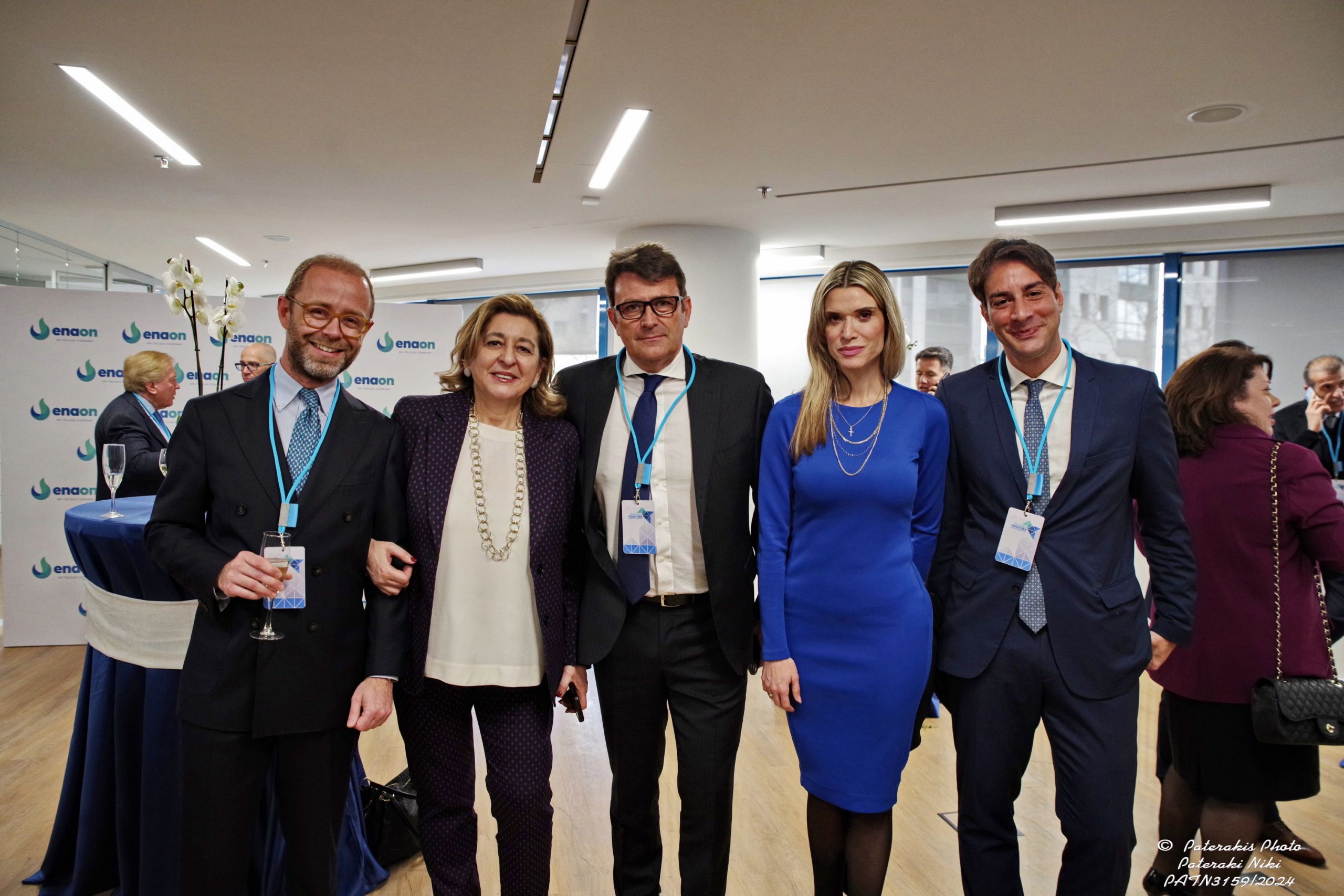 Cinque persone sorridenti in abito formale con badge in un evento aziendale con un pannello con i lighi Enaon sullo sfondo.