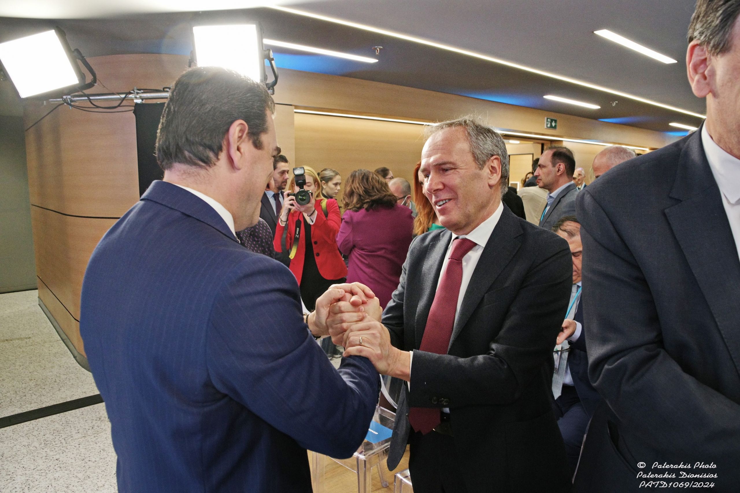 Paolo Gallo e un altro uomo in abito si stringono le mani sorridenti in un ambiente affollato e illuminato.