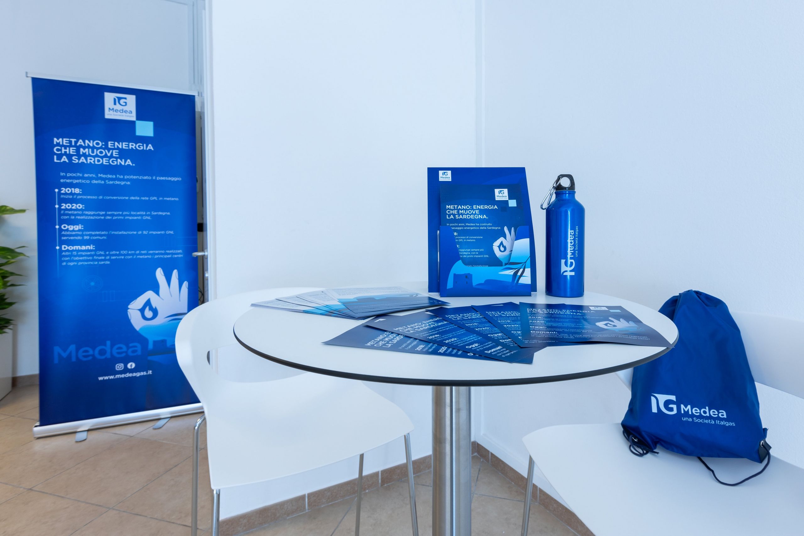 Angolo informativo Medea Italgas con depliant, borraccia e zaino su tavolo e roll-up accanto.
