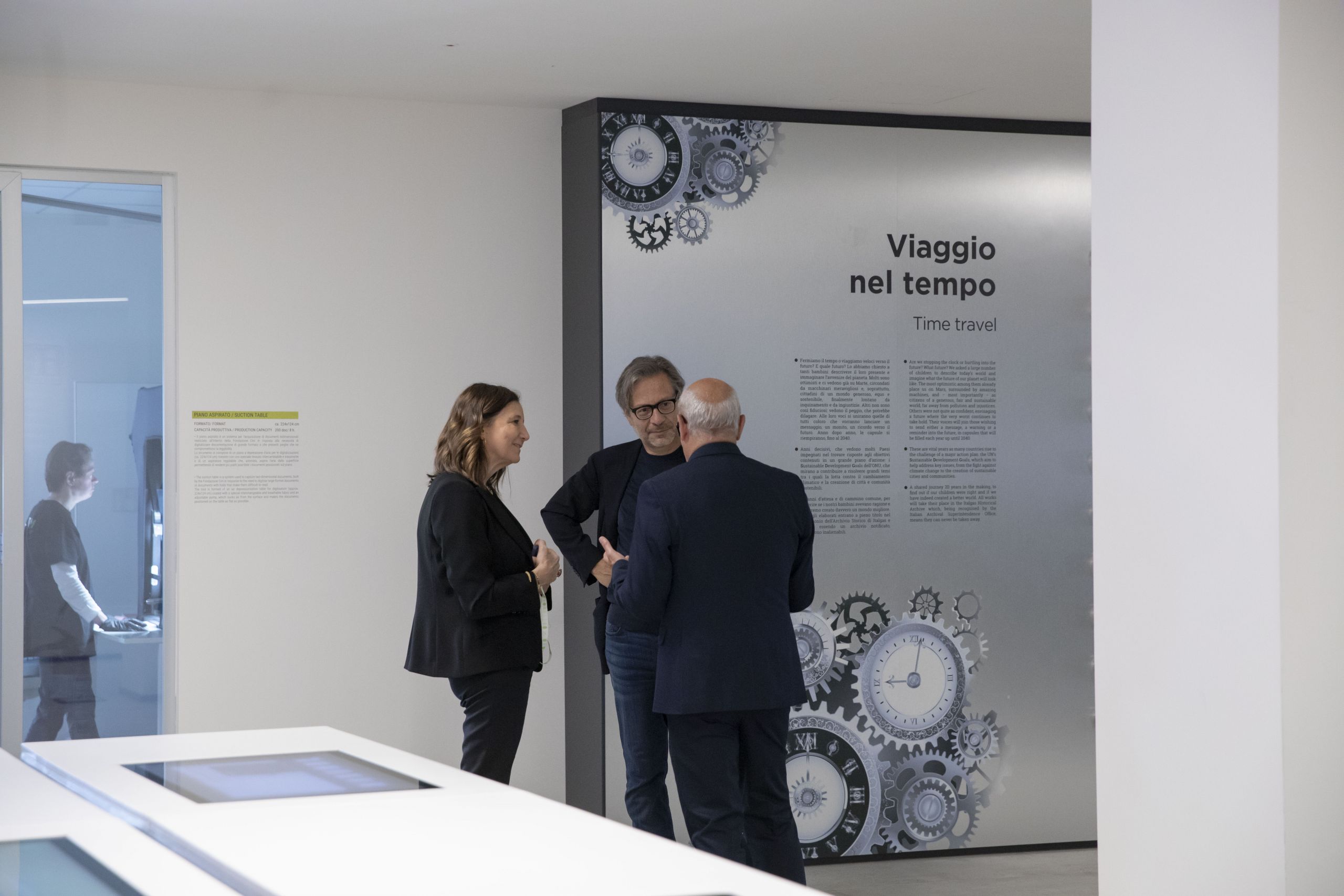 Recalcati conversa con altre figure davanti a un pannello espositivo su "Viaggio nel tempo" in uno spazio di Heritage lab.