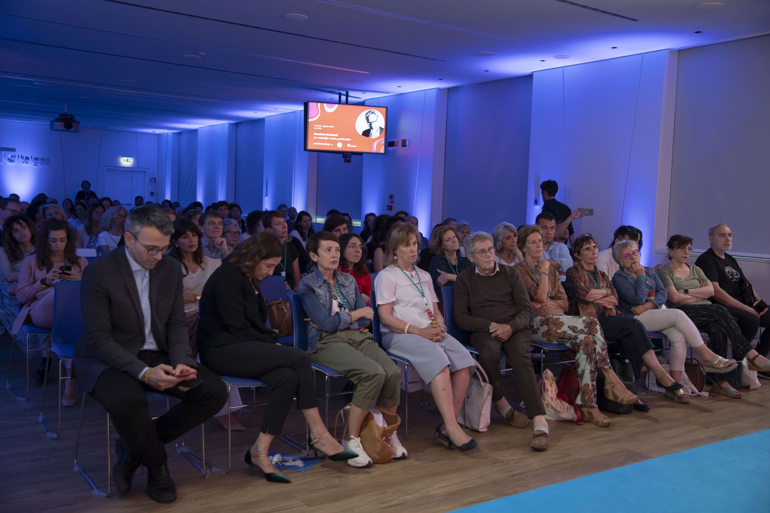 Pubblico attento all'evento Heritage lab in una sala conferenze con luci soffuse e schermo al fondo.