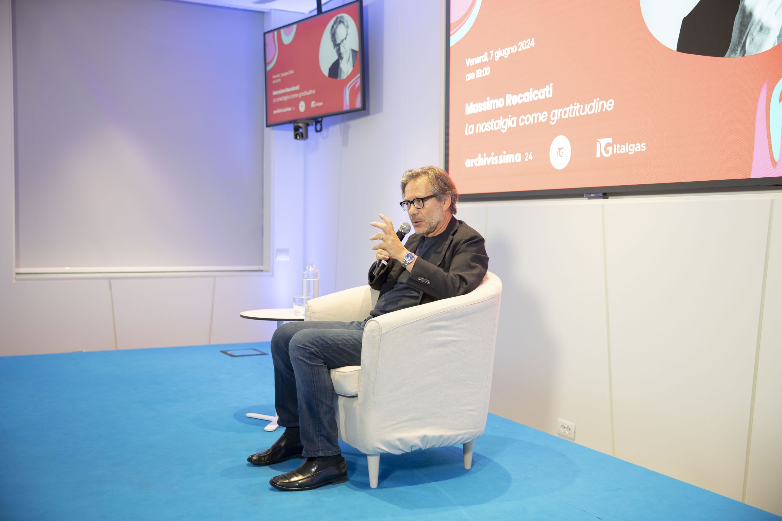 Recalcati seduto su poltrona bianca mentre parla al microfono in evento Heritage lab. Schermo con dettagli evento sullo sfondo.