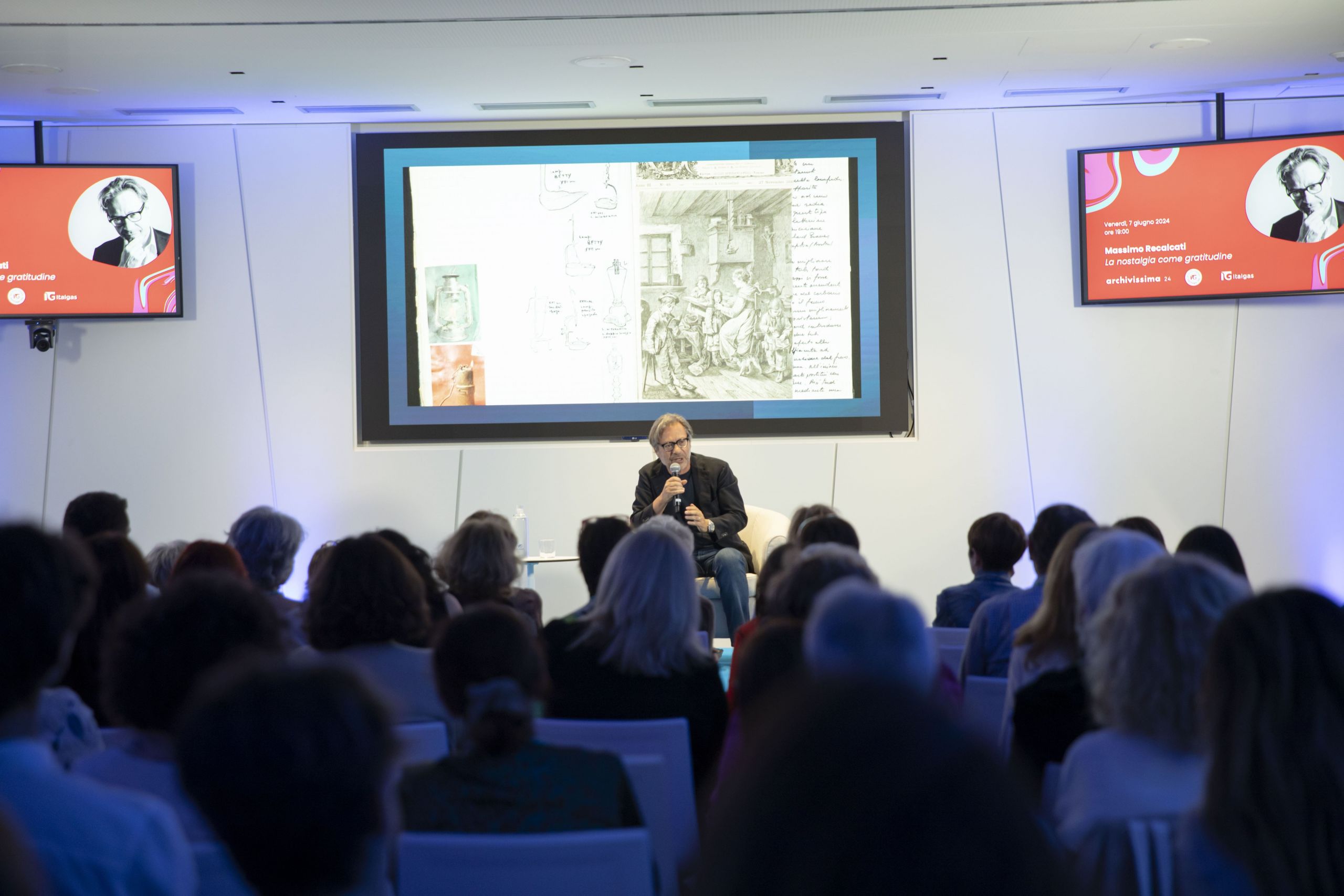 Conferenza all'evento Heritage lab, relatore con microfono, pubblico attento, slide con arte sullo sfondo.