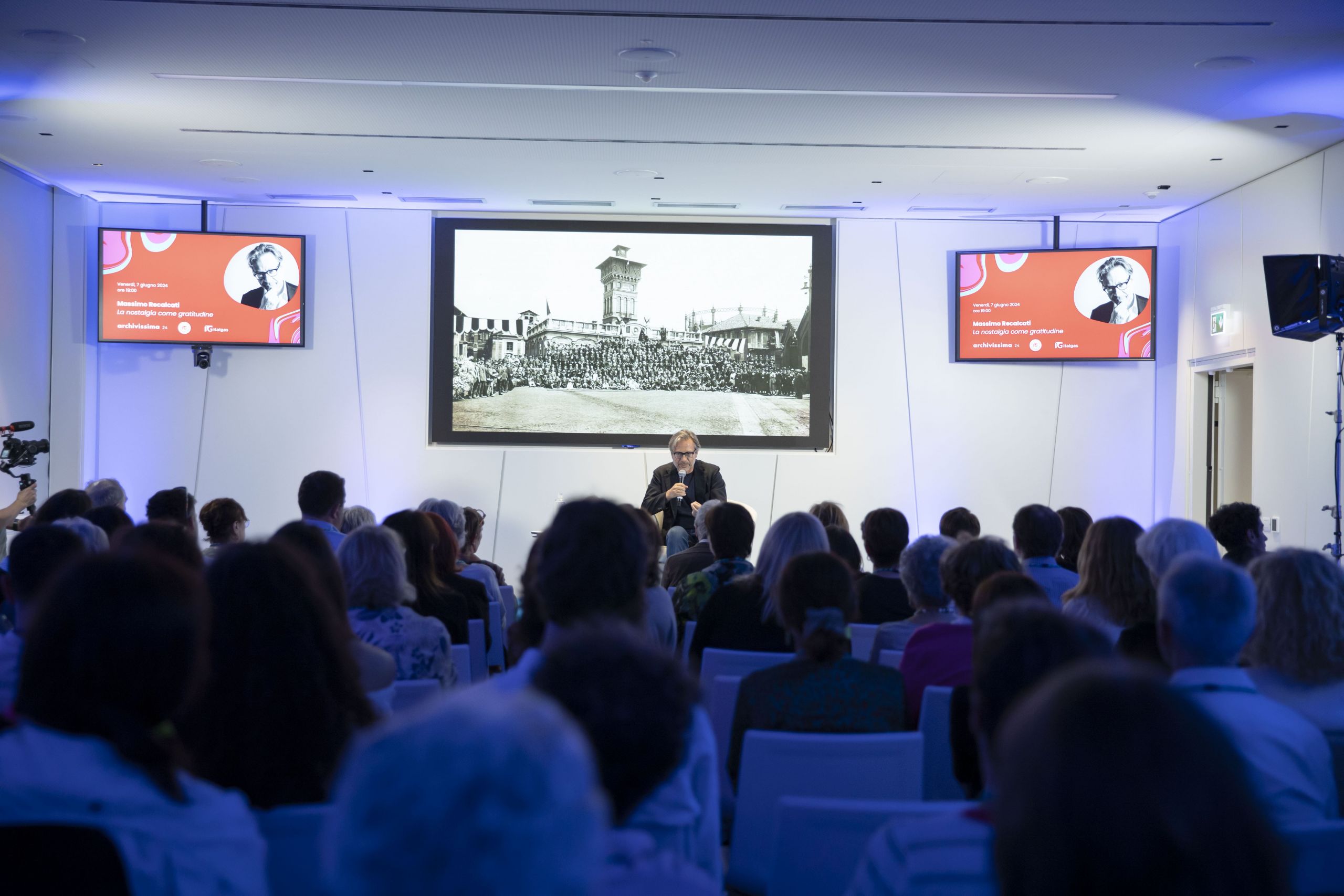 Pubblico seduto ascolta relatore al Heritage lab evento, monitor mostra immagine d'epoca.