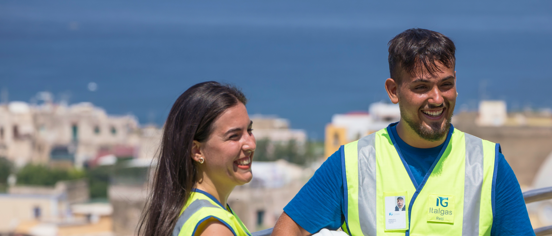 Due lavoratori Italgas sorridenti in giubbotto riflettente e paesaggio con mare sfondo.