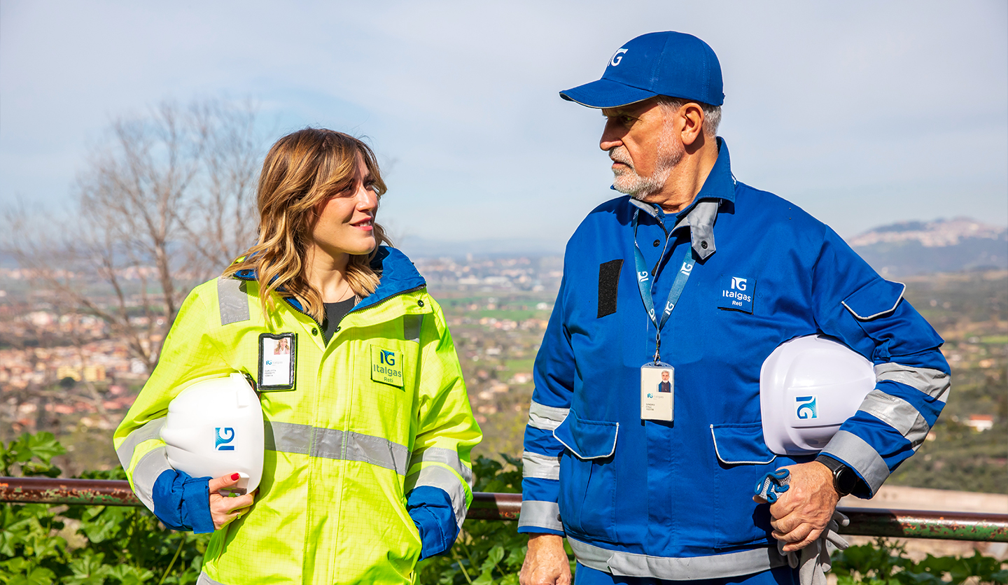 Due dipendenti Italgas un uomo e una donna in divise da lavoro e caschi protettivi conversano all'aperto.